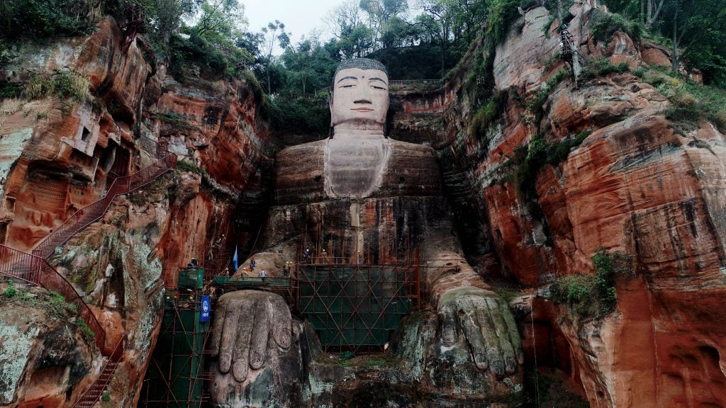 the gigantic leshan buddha statue sits embedded between two rock faces, with vegetation growing on and around it. its pose is serene, with its hands placed peacefully on its knees and a neutral expression on its face.