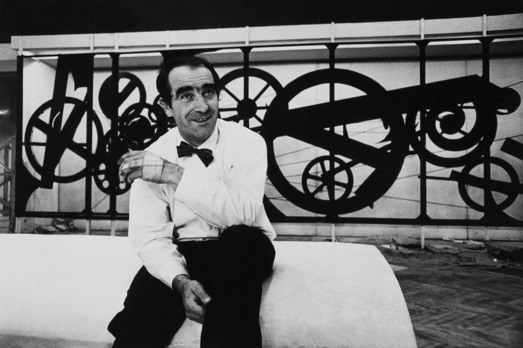 a man in a bow tie sits in front of a geared sculpture 