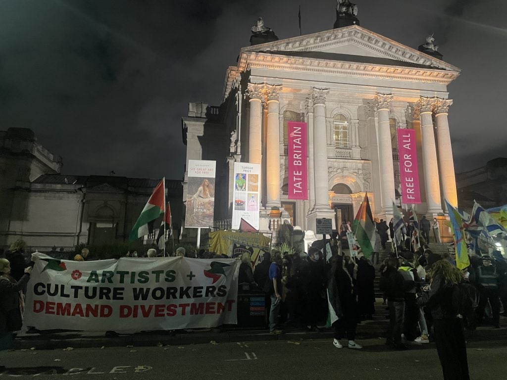 a crowd of people holding signs stand out front of a large lit up neoclassical building at night