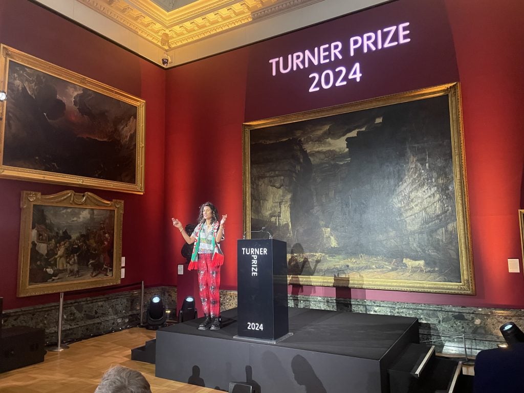 a woman stands on stage near a podium in a large room with red walls hung with paintings