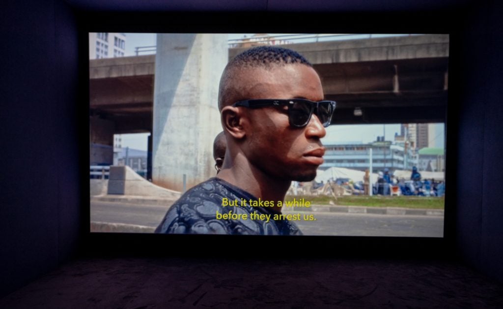 a photo of an installation of a film in a dark place, a still on the screen shows a man wearing sunglasses in an urban setting with the subtitles reading 'but it takes a while before they arrest us.'