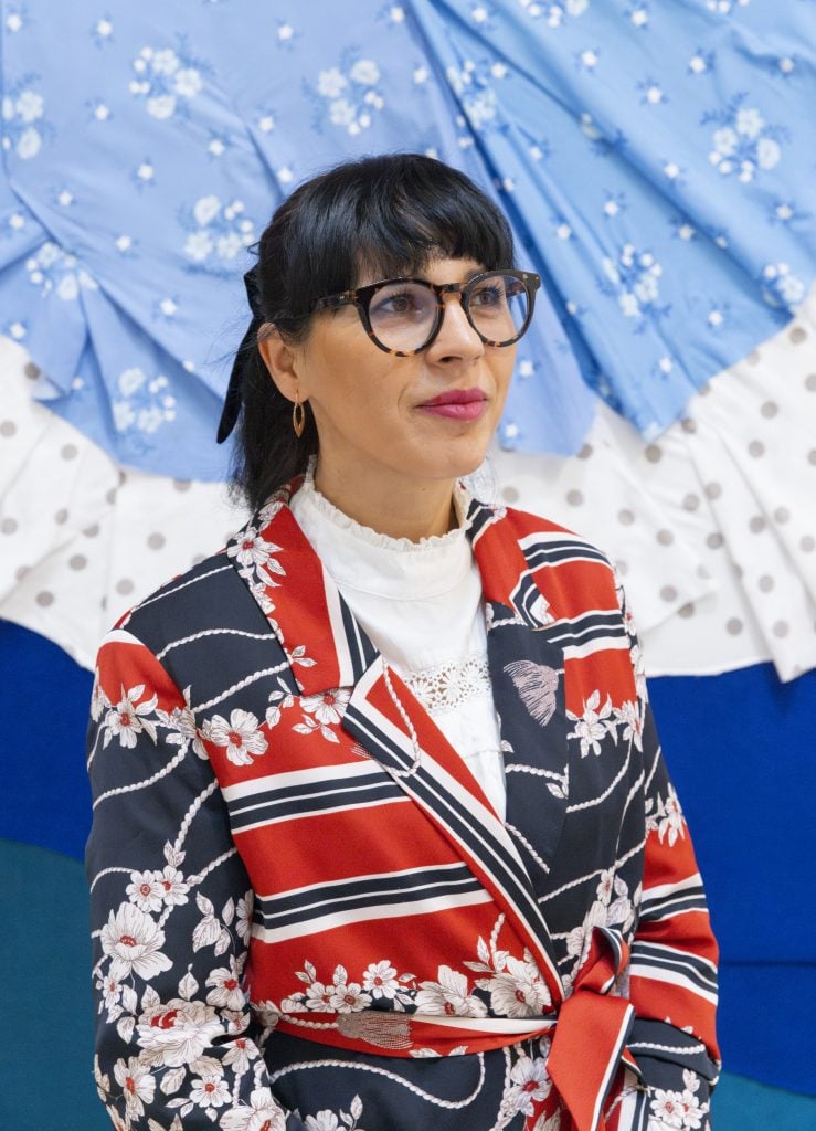 Małgorzata Mirga-Tas at Tate St. Ives, (2024). She is a young woman with dark bangs and her hair in a ponytail, wearing a striped red and black blazer with a flower print, standing in front of a blue and white textile collage.