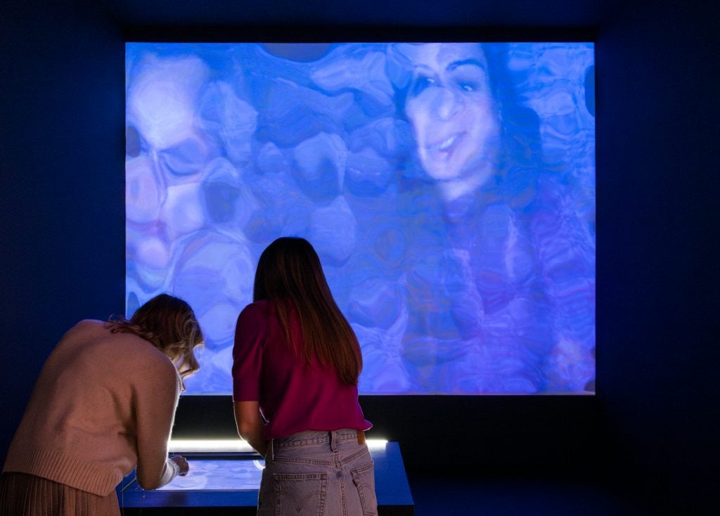 two figures from behind are seen bending down and inspecting and doing things on a screen on a table, they are facing a screen with a blurry purple and blue image that includes a woman's face amid the blurriness