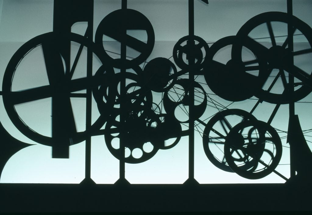 a geared Jean Tinguely art piece is backlit dramatically 