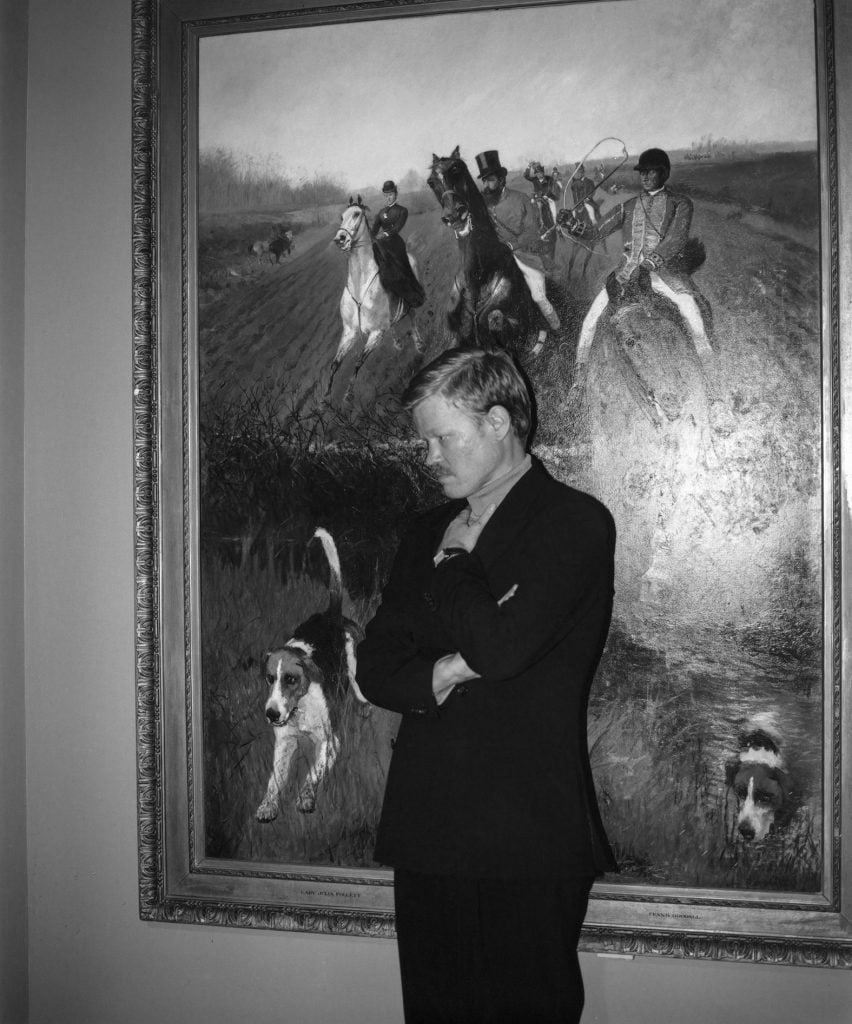 A man in formal attire poses in front of a framed hunting scene with dogs.