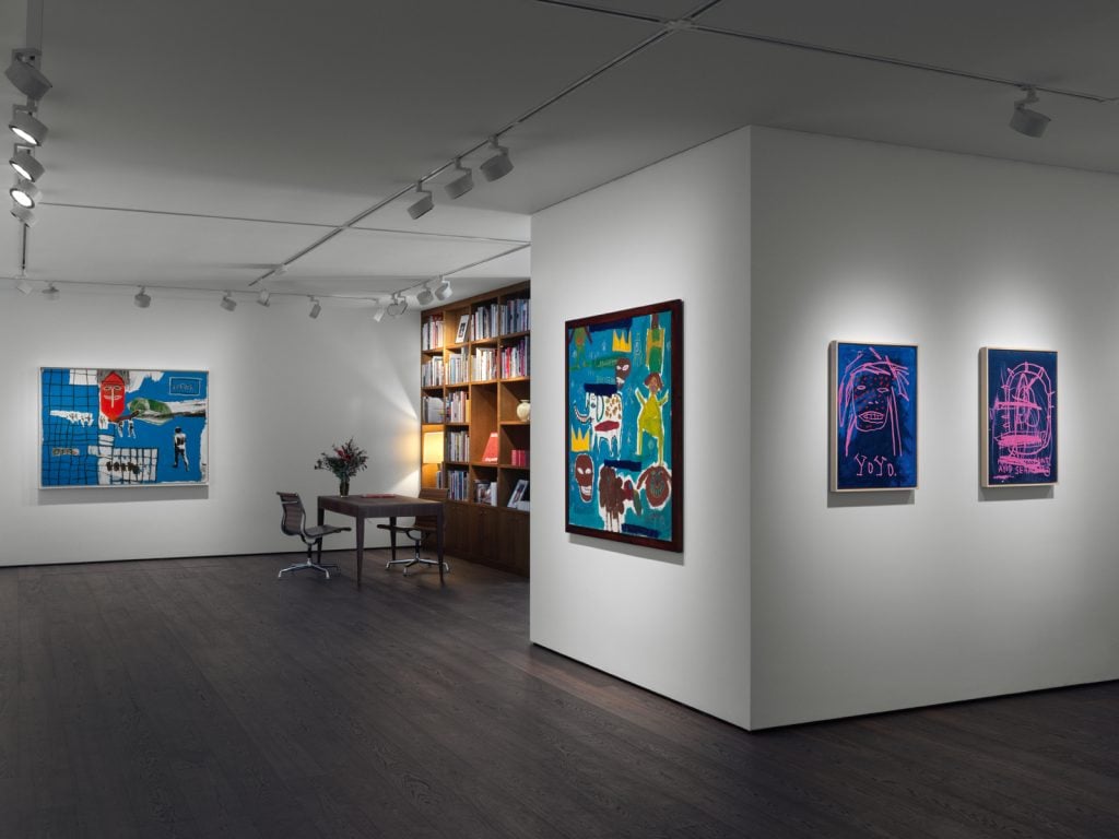 A photograph of a Basquiat paintings hanging throughout a white walled gallery, with a bookshelf, desk and chair nestled on the other side of a wall.