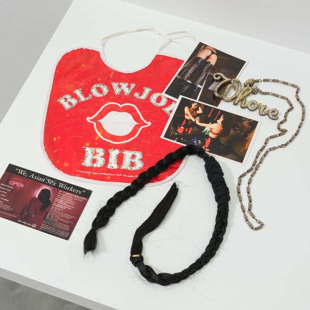 Objects on a white surface related to sex work including a red bib, photographs, postcard, necklace, and hair braid. 