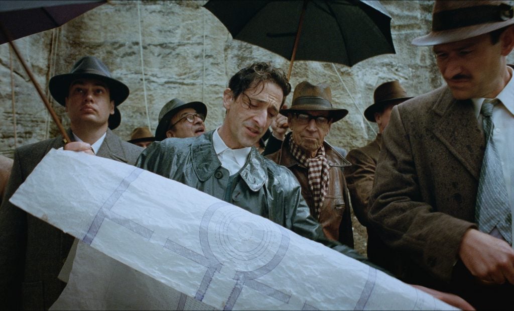 A man holding up design plans for a building, while surrounded by a crowd of other men