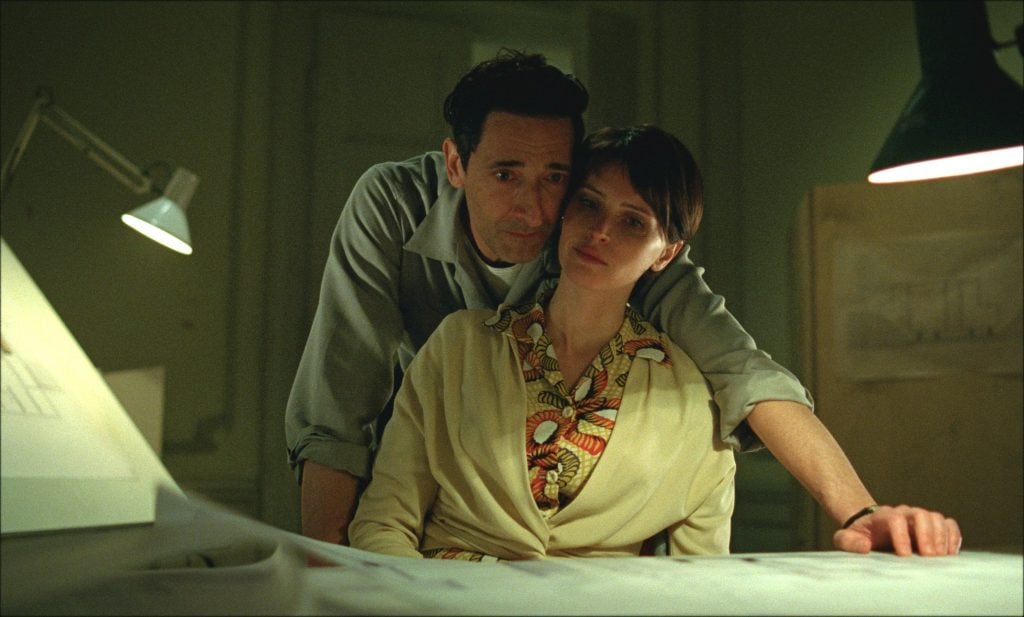 Actor Adrien Brody affectionately looks over a seated Felicity Jones's shoulder in the film The Brutalist