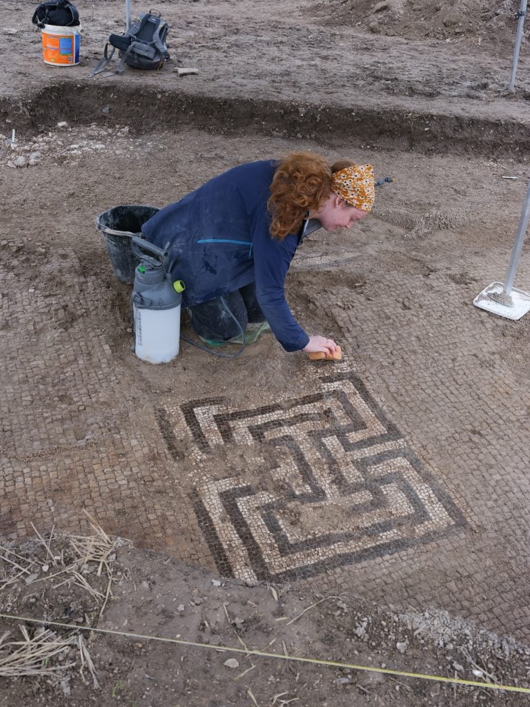 A photograph of an archaeologist brushing a Roman mosaic emerging from the earth.