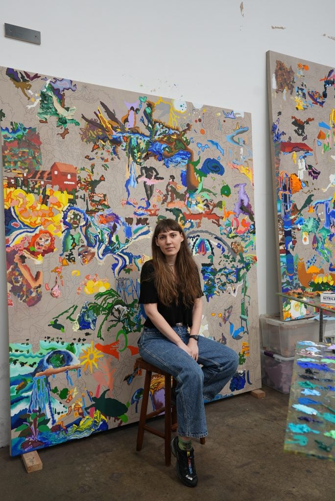 Portrait of the artist Cindy Phenix sitting on a stool in front of one of her paintings wearing blue jeans and a black t-shirt with long loose brown hair, artist included in shows to see list.