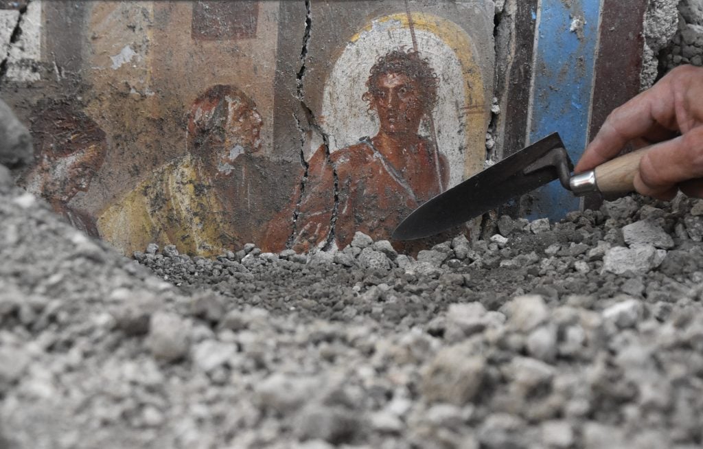 A fresco being excavated by a hand with a trowel, half-covered in rocks.