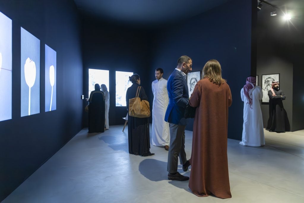  Visitors interact at a modern art exhibition at Diriyah Art Futures with illuminated digital screens and framed artworks in a dimly lit gallery space, creating an immersive viewing experience.