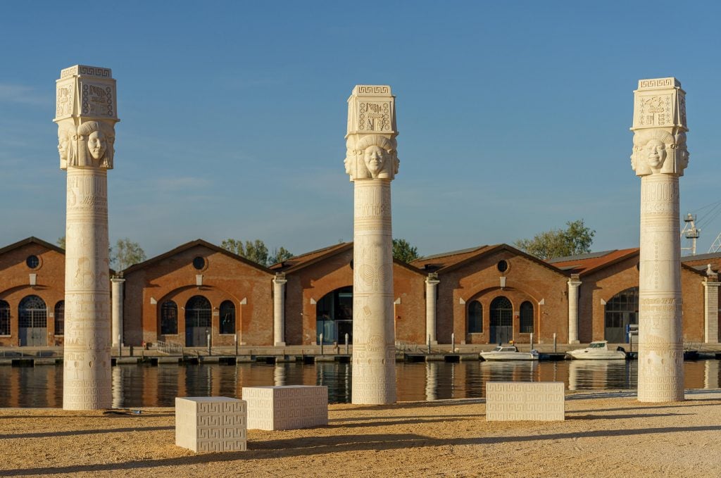 an artist's modern update of ancient Egyptian columns has contemporary black faces and iconography and is in front of a body of water and an industrial building 