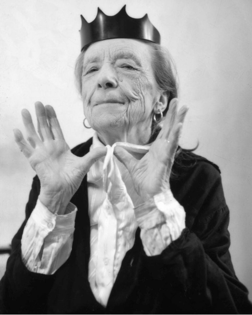 Portrait of Louise Bourgeois, wearing a party hat shaped like a crown, with her hands splayed in front of her