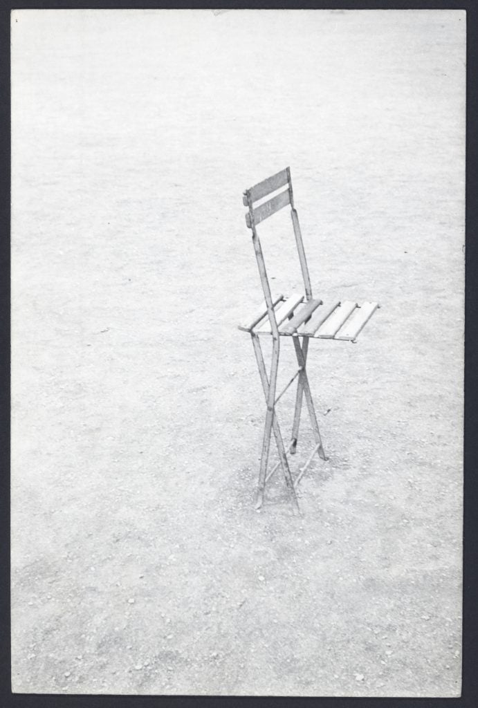 A black and white photograph of a folding chair on bare dirt ground.