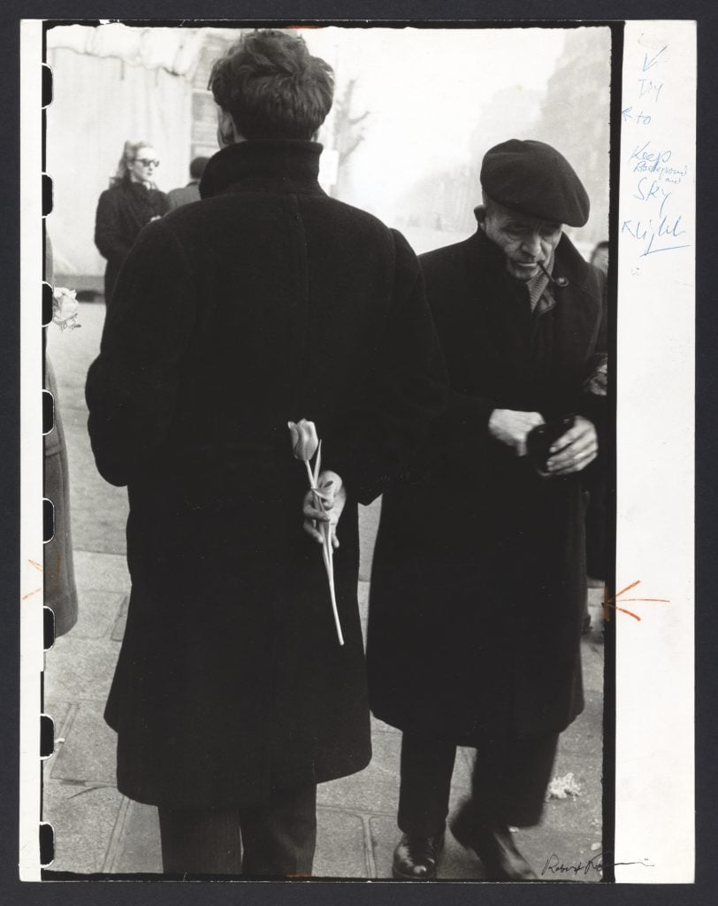 A black and white photograph of two men in black coats and hats, one facing away with a single tulip clutched behind his back.