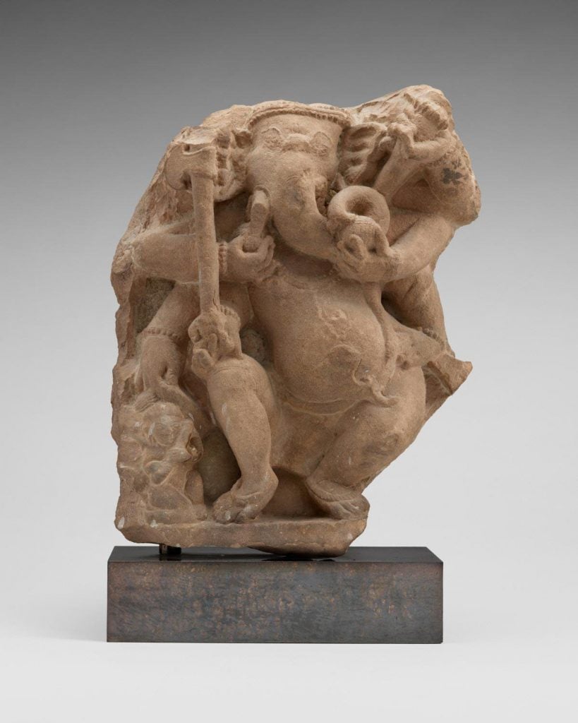 small brown statue on a small pedestal of a humanoid creature with an elephant for a head with multiple arms holding a stick against a lit grey background