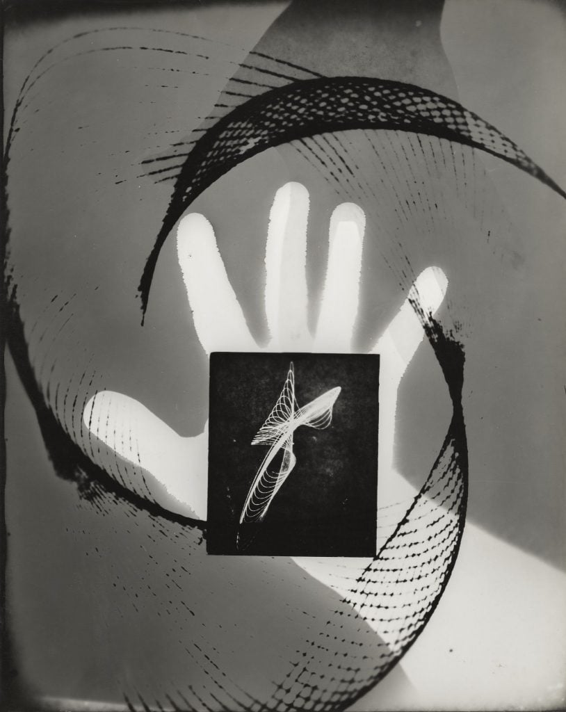 A Surrealist black and white photograph depicting an extended white hand with black lines swirling around it, and a white abstract form within a black square atop its palm, all on a gray background.