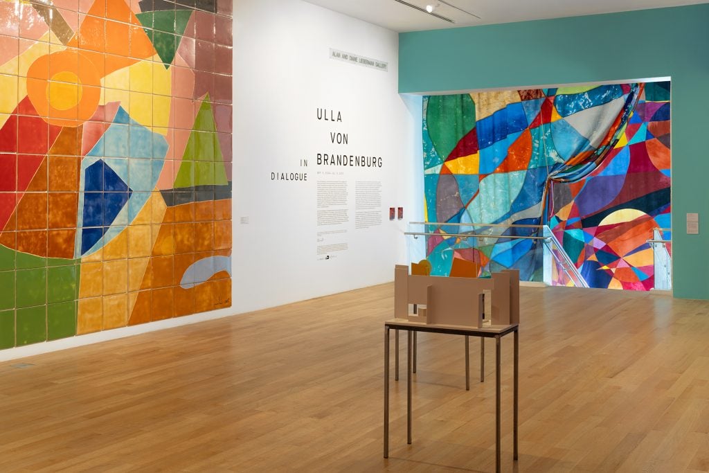 A photograph of Ulla von Brandenburg's show on view at The Bass, with Etel Adnan's cermaic mural to the left, a colorful geometric abstraction by von Brandenburg grounded in a teal wall to the right, and a small sculpture on a table atop the wood floor before them both.